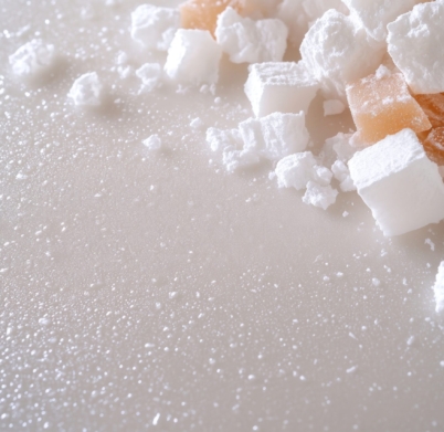 This close-up captures a scattering of sugar cubes, some dusted with what appears to be confectioner's sugar, alongside translucent amber-colored pieces on a reflective white surface. The composition suggests a sweet, possibly culinary theme, emphasizing the textures of the granulated sugar and the smooth surface beneath. The lighting is soft and diffused, enhancing the visual appeal for possible usage in food-related content, baking advertisements, or dessert presentations, creating a palatable and inviting image. The focus on texture and color also lends itself well to abstract or minimalist backgrounds.