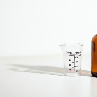 An amber glass bottle with a white cap sits next to a clear plastic measuring cup with dosage markings on a clean white surface.