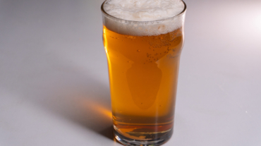 A vertical studio shot of a full pint glass filled with golden amber beer. The beverage features a thick, white, frothy head of foam and visible carbonation bubbles rising through the liquid. The glass is a classic nonic pint shape, casting a distinct shadow to the left on a plain, light grey background. The composition includes copy space on the left side.