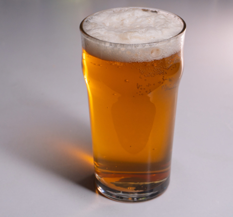 A vertical studio shot of a full pint glass filled with golden amber beer. The beverage features a thick, white, frothy head of foam and visible carbonation bubbles rising through the liquid. The glass is a classic nonic pint shape, casting a distinct shadow to the left on a plain, light grey background. The composition includes copy space on the left side.