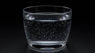 A clear glass filled with sparkling water sits against a stark black backdrop. The drink bubbles with effervescence, its transparent liquid filled with numerous tiny bubbles. The glass is simple in design, with smooth, curved lines. The scene is illuminated to highlight the texture and clarity of the water. This image could be used for advertisements, articles, or other projects related to hydration, beverages, or the refreshment industry. It evokes feelings of cleanliness and thirst-quenching.