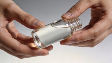 View shows pair of bare hands grasping clear glass dropper bottle filled with thick transparent fluid, soft lighting creating glossy reflections on smooth surfaces against neutral gray backdrop