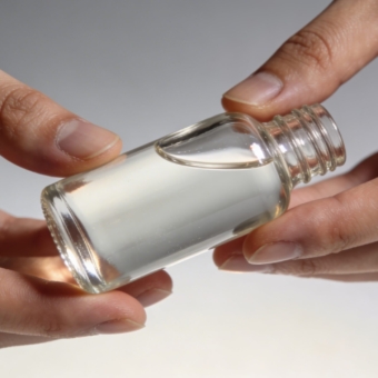 View shows pair of bare hands grasping clear glass dropper bottle filled with thick transparent fluid, soft lighting creating glossy reflections on smooth surfaces against neutral gray backdrop
