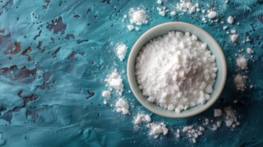 Overhead shot showcases a small bowl filled with a generous amount of white powder against a textured blue backdrop. The fine powder also scatters around the bowl, creating a visually appealing contrast with the background's rough texture. The scene is illuminated, accentuating the details of the powder and the textured surface. The image would be suitable for use in articles or advertisements related to baking ingredients, food preparation, or healthy eating. It could also be used for illustrating concepts related to purity or cleanliness.