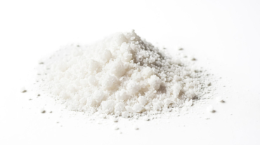 Close-up shot of white salt crystals on white background. The salt crystals are piled up in a small heap and are illuminated by bright light. The background is blurred and out of focus.