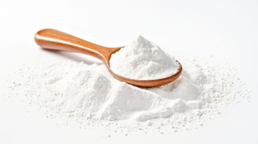 A wooden spoon filled with white powder rests on a white background, surrounded by a scattering of the same powder.